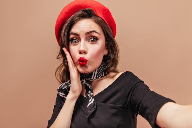 Fille aux yeux verts aux cheveux noirs avec des lèvres rouges regarde la caméra avec surprise et prend selfie sur fond beige.