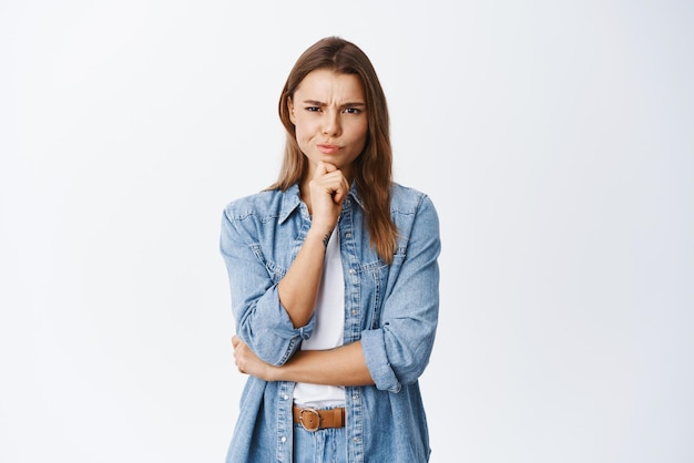 Fille ayant des doutes Femme suspecte fronçant les sourcils et regardant la caméra réfléchie hésitant à propos de l'accord debout pensif sur fond blanc