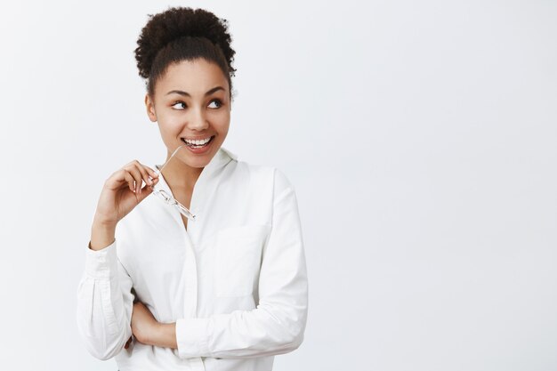 Fille ayant l'intention d'aller dîner avec un homme charmant du prochain bureau. Intrigué belle femme africaine en chemise blanche, regardant à droite et souriant largement, mordant le bord de lunettes de curiosité