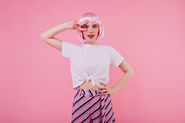 Fille bien faite extatique en tenue à la mode posant avec une expression de drôle de visage sur le mur rose. modèle féminin insouciant en t-shirt blanc et Pérou brillant