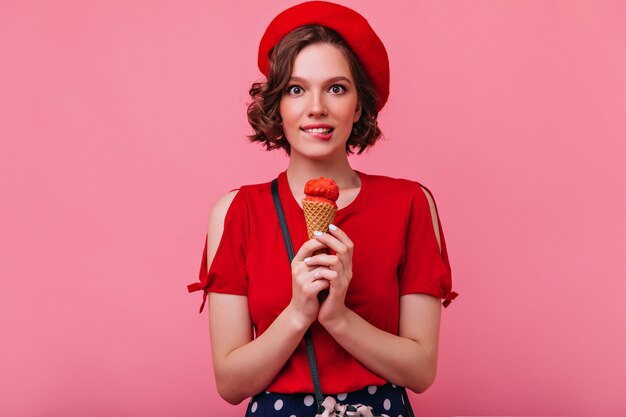 Fille blanche glamour en chemisier rouge, manger de la crème glacée. Modèle féminin français de bonne humeur debout avec dessert.