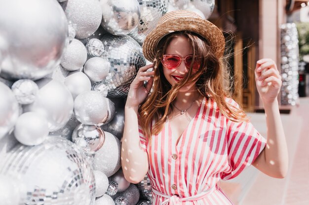 Photo gratuite fille blanche insouciante en robe rayée posant près de boules scintillantes. portrait en plein air d'un modèle féminin adorable en chapeau de paille se détendre en été.