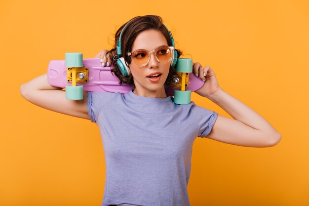 Fille blanche inspirée dans des lunettes de soleil élégantes posant avec longboard coloré. Photo intérieure d'un modèle féminin intéressé dans de gros écouteurs à la mode.