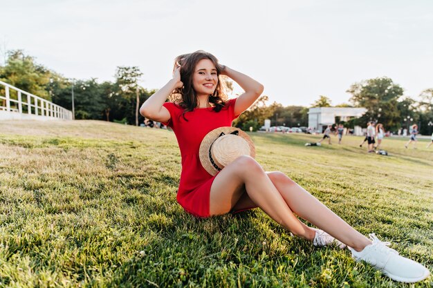 Fille blanche positive assise sur l'herbe et jouant avec ses cheveux bruns. Joyeuse dame en riant en robe rouge profitant du matin du week-end dans le parc.