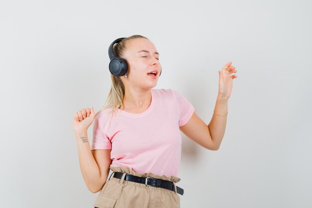 Fille blonde appréciant la musique avec des écouteurs en t-shirt, pantalon et à la fringante. vue de face.