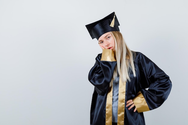 Fille blonde appuyée sur la joue de la paume tout en tenant la main sur la taille en robe de graduation et casquette et à la charmante