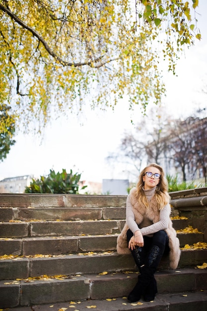 Fille blonde au manteau de fourrure et lunettes assis sur les escaliers avec des feuilles jaunes