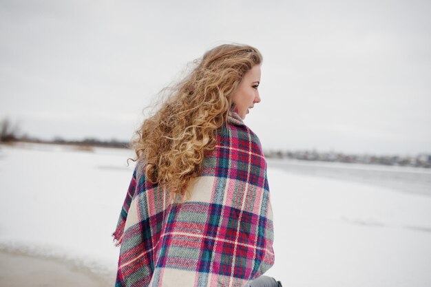 Fille blonde bouclée en plaid à carreaux contre le lac gelé au jour d'hiver