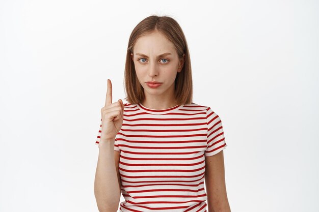 Fille blonde bouleversée pointant le doigt vers le haut, fronçant les sourcils mécontent, n'aimant pas qch, exprime son opinion négative, chose injuste, debout sur fond blanc en t-shirt