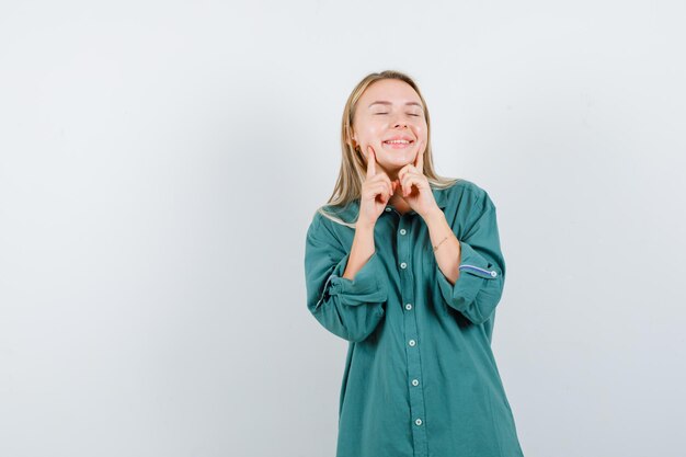 Fille blonde en chemisier vert tenant l'index près de la bouche, forçant un sourire et l'air heureux