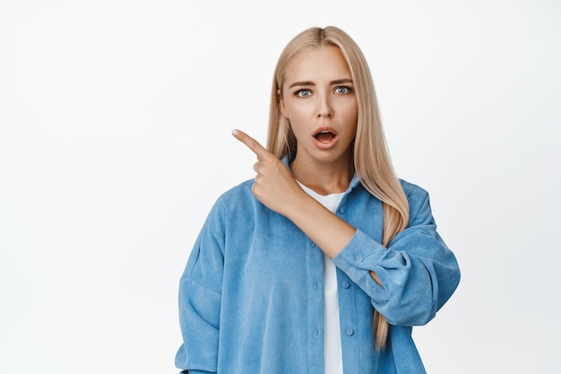 Fille blonde choquée regardant avec admiration avec la mâchoire tombée pointant le doigt dans le coin supérieur gauche montrant le fond blanc de la bannière du logo