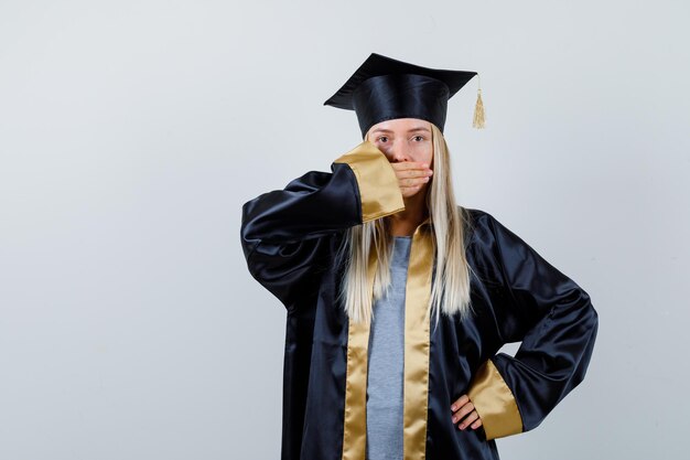 Fille blonde couvrant la bouche avec la main tout en tenant la main sur la taille dans une robe de graduation et une casquette et l'air surpris.