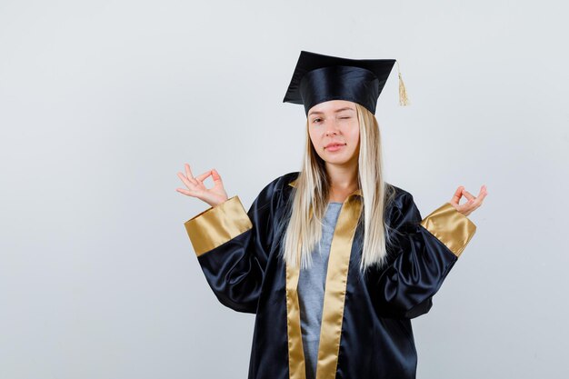 Fille blonde debout dans une pose de méditation, un clin d'œil dans une robe de graduation et une casquette et l'air heureux