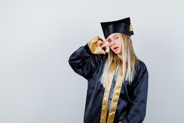 Fille blonde debout tout droit, montrant le signe v sur l'œil et posant à la caméra en robe de graduation et casquette et l'air mignon.