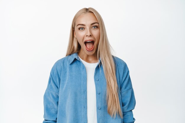 Une fille blonde drôle ouvre grand la bouche, crie et a l'air idiot, debout sur du blanc.