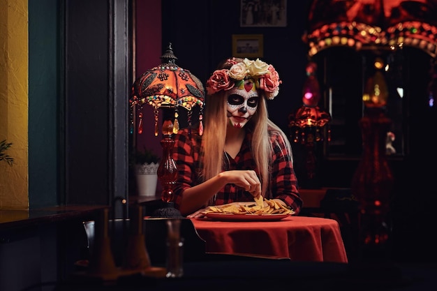 Fille blonde effrayante avec un maquillage mort-vivant dans une couronne de fleurs mangeant des nachos dans un restaurant mexicain. Concept d'Halloween et de Muertos.