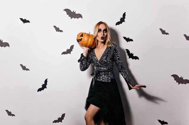 Fille blonde élégante en robe noire posant à la fête de la sorcière. Modèle féminin élégant avec un maquillage sombre tenant une grosse citrouille d'halloween.