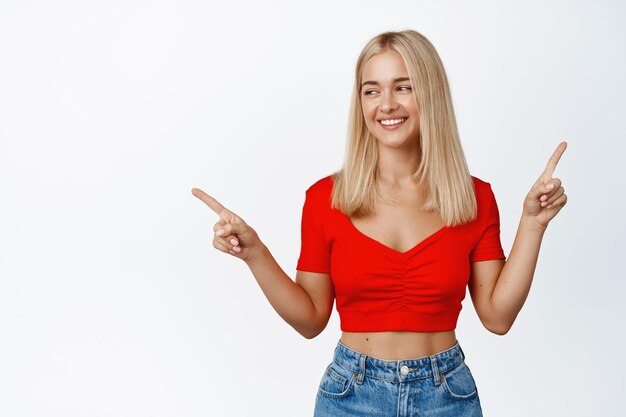 Une fille blonde élégante sourit montre deux façons de se diriger vers le côté et a l'air heureuse de choisir un produit debout sur fond blanc