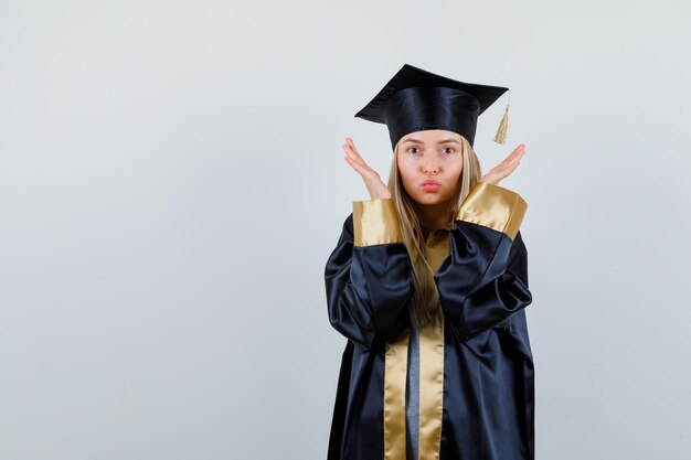 Fille blonde étirant les mains près du visage, envoyant des baisers dans une robe de graduation et une casquette et ayant l'air heureuse.