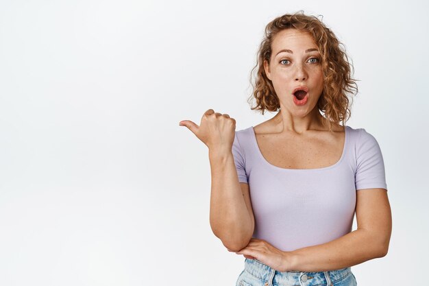 Une fille blonde étonnée laisse tomber la mâchoire, pointant le doigt vers la gauche et regardant avec admiration, réagissant à une annonce impressionnante, montrant la vente, debout sur fond blanc