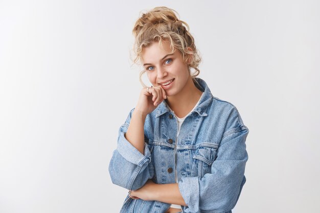 Fille blonde européenne élégante à la mode à la recherche d'une caméra curieuse souriante intriguée écouter une suggestion intéressante souriante pensant avoir une bonne idée, debout mur blanc heureux