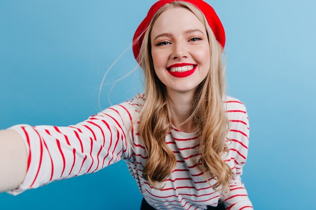 Photo gratuite fille blonde excitée en béret prenant selfie sur mur bleu. insouciante jeune femme en chemise rayée.