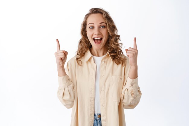 Fille blonde excitée haletant, pointant les doigts vers le haut et criant de joie, racontant de grandes nouvelles, l'air étonné, debout sur un mur blanc