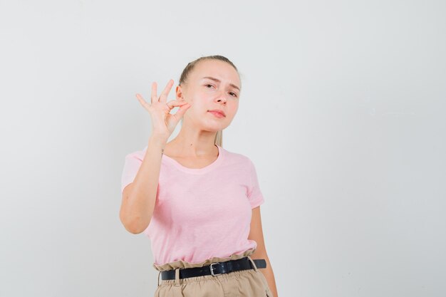 Fille blonde faisant un geste ok en t-shirt, pantalon et à la vue de face, confiant.