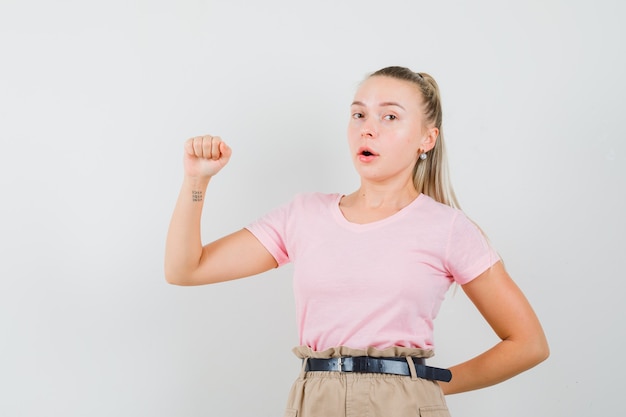 Fille blonde faisant semblant de tenir ou de montrer quelque chose en t-shirt, pantalon, vue de face.