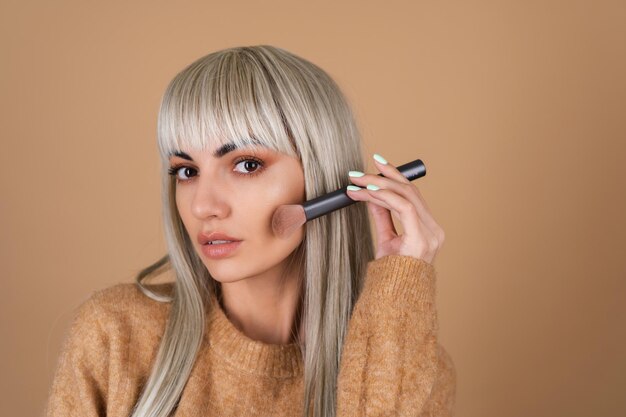 Une fille blonde avec une frange et un maquillage de jour brun tient un pinceau pour le contouring et le surligneur