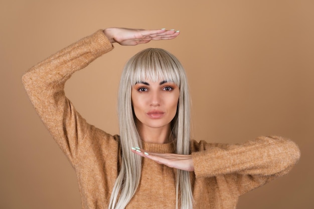 Une fille blonde avec une frange et un maquillage de jour marron dans un pull sur fond beige tire ses mains au-dessus de sa tête et sous son menton