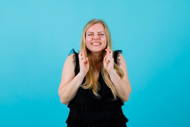 La fille blonde heureuse croise des doigts sur le fond bleu