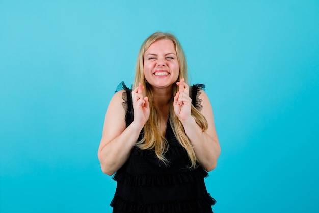 La fille blonde heureuse croise des doigts sur le fond bleu