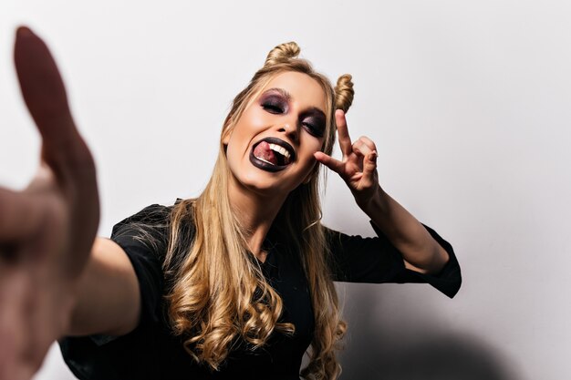 Fille blonde heureuse avec des lèvres noires faisant selfie à halloween. Incroyable jeune sorcière drôle posant sur un mur blanc.