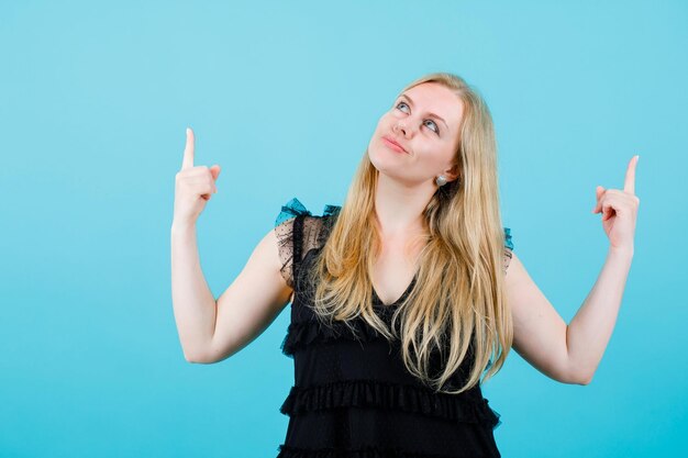 Une fille blonde heureuse regarde et pointe vers le haut avec les index sur fond bleu