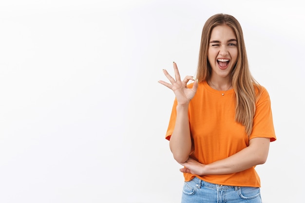 Une fille blonde impertinente montre un signe d'accord, un clin d'œil et un sourire, assure que vous aimerez cela, le meilleur choix jamais fait, garantit la meilleure qualité, satisfaite après avoir passé les cours, e-learning, confiante quant à la décision