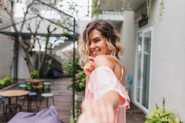 Fille blonde ludique regardant par-dessus l'épaule avec un sourire timide. Photo extérieure d'un modèle féminin caucasien effrayant s'amusant dans un café de rue.