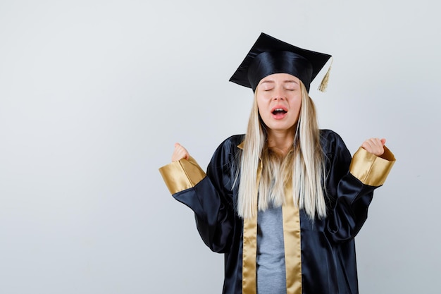 Photo gratuite fille blonde montrant le geste du gagnant en robe de graduation et casquette