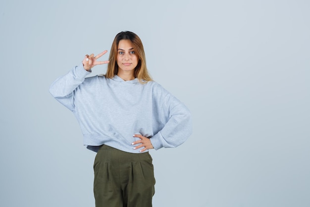 Fille blonde montrant un geste de paix et tenant la main sur la taille en sweat-shirt et pantalon bleu vert olive et l'air radieuse, vue de face.