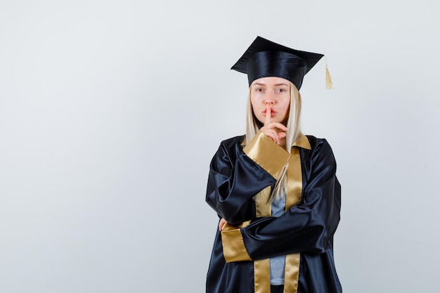 Fille blonde montrant un geste de silence en robe de graduation et casquette et semblant sérieuse.