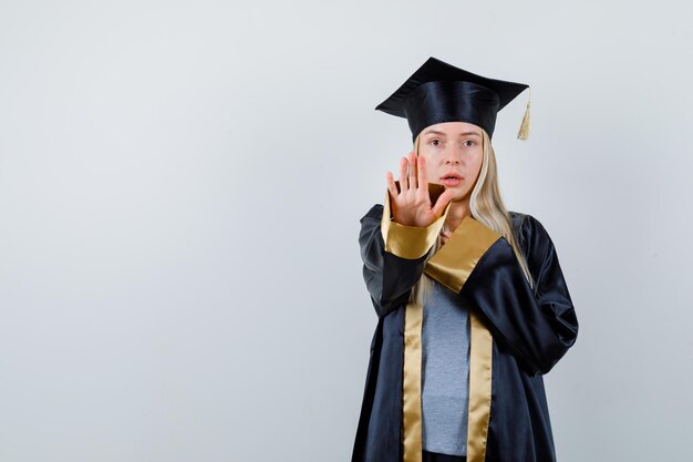 Fille blonde montrant un panneau d'arrêt, reposant la main sur la poitrine en robe de graduation et casquette et semblant sérieuse.