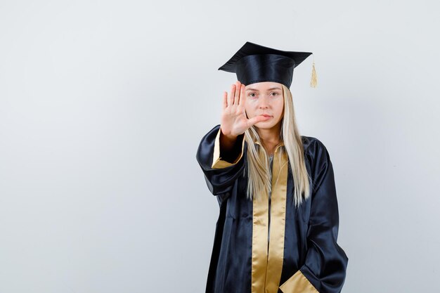 Fille blonde montrant le panneau d'arrêt en robe de graduation et casquette et à la recherche de sérieux.