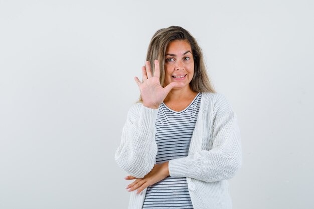 Fille blonde montrant un panneau d'arrêt tout en tenant une main sous le coude en t-shirt rayé, cardigan blanc et pantalon en jean et à la recherche de plaisir. vue de face.