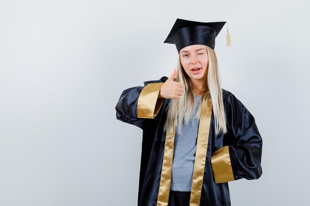 Fille blonde montrant le pouce vers le haut tout en tenant la main sur la taille, un clin d'œil en robe de graduation et casquette