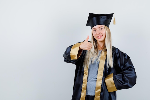 Fille blonde montrant le pouce vers le haut tout en tenant la main sur la taille dans une robe de graduation et une casquette et l'air heureux