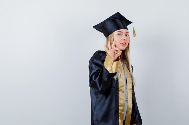 Fille blonde montrant un signe ok dans la robe de graduation et la casquette et ayant l'air heureux