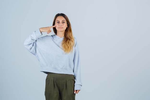 Fille blonde pointant sur elle-même avec l'index en sweat-shirt et pantalon bleu vert olive et l'air sérieux. vue de face.