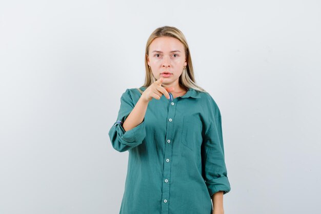 Fille blonde pointant vers la caméra avec l'index en blouse verte et à la recherche d'enchantement