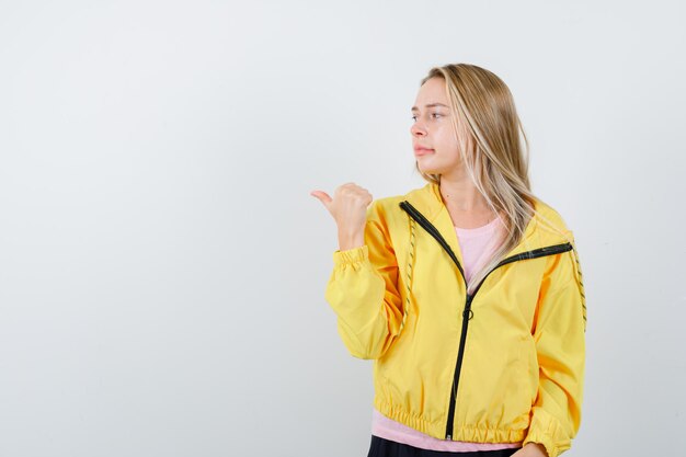 Fille blonde pointant vers la gauche en t-shirt rose et veste jaune et l'air heureux.