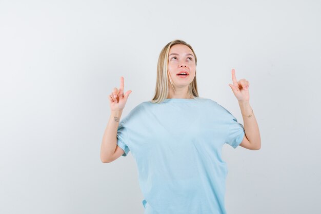 Fille blonde pointant vers le haut avec l'index en t-shirt bleu et à la recherche de bonheur. vue de face.